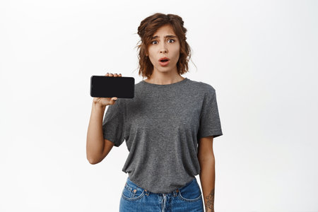 Shocked and concerned young woman gasps, shows mobile phone screen, horizontal interface on smartphone application, standing over white backgroundの写真素材