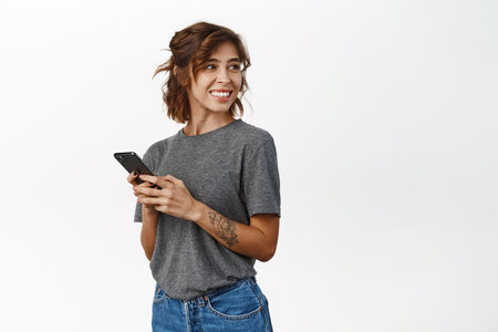 Smiling authentic girl using smartphone app, chatting or ordering, holding mobile phone, standing against white backgroundの写真素材