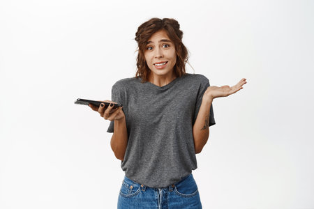 Young confused woman with smartphone, shrugging shoulders and holding mobile phone, dont know, being indecisive, standing over white backgroundの写真素材