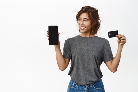 Portrait of cute girl with phone, looks and shows smartphone screen, holds black credit card, white backgroundの写真素材