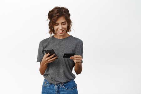 Online shopping. Smiling young woman using mobile phone and credit card to pay, make purchase, order or register in web store, standing over white backgroundの写真素材