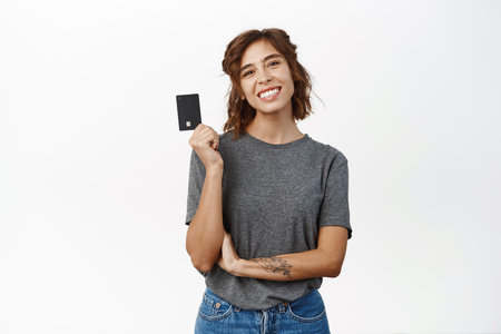 Portrait of beautiful young woman showing credit card, recommending bank, contactless shopping or discounts in store, time to shop, standing over white backgroundの写真素材