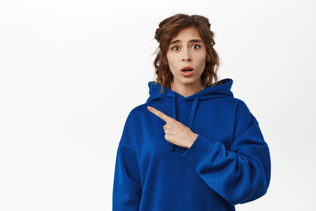 Worried insecure young woman pointing left at empty space, looking concerned at camera, standing over white backgroundの写真素材