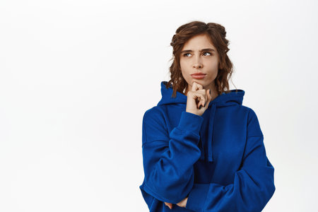 Portrait of girl thinking, looking aside and deciding, making choice, standing in blue hoodie, gazing at upper left corner, empty copy space, white backgroundの写真素材