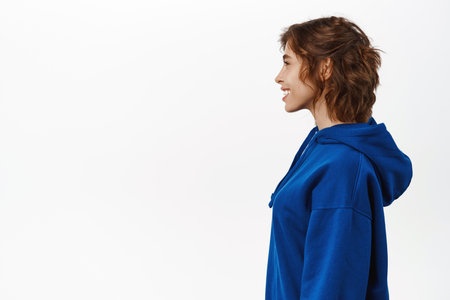 Profile portrait of young woman with curly hairstyle, wearing casual blue hoodie, smiling and looking to the left, standing over white backgroundの写真素材