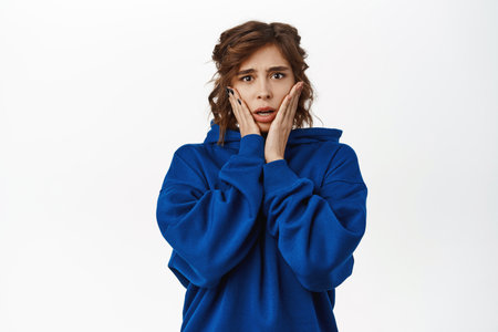 Shocked young woman, horrified, looking scared or worried, standing in blue hoodie over white backgroundの写真素材