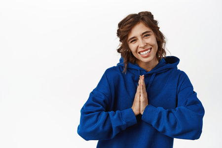 Thank you. Smiling cute brunette girl showing namaste, gratitude gesture, thanking, bowing politely, standing in blue hoodie over white backgroundの写真素材