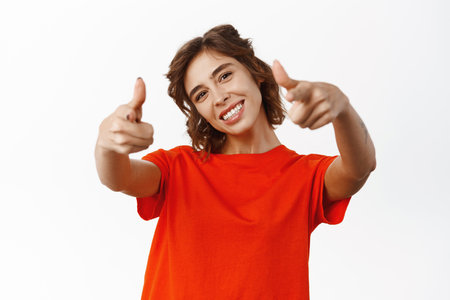 Portrait of beautiful bright woman pointing fingers at camera and smiling pleased, congratulating, praise you, inviting to store, standing over white backgroundの写真素材