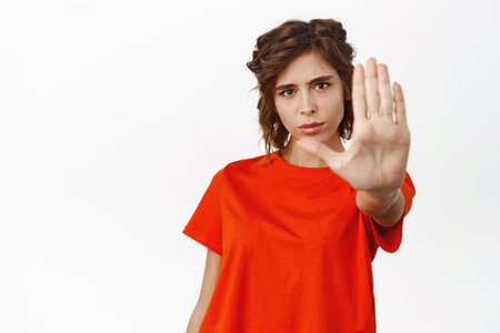 Image of serious young woman showing block, stop gesture, extending arm palm and saying no, rejecting, declining smth, stay back sign, standing over white backgroundの写真素材