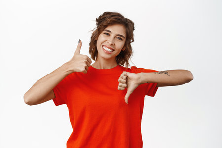Portrait of brunette female model showing thumbs up and thumbs down, like or dislike gesture, deciding, choosing, standing over white backgroundの写真素材