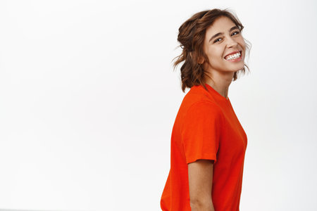 Portrait of attractive young hispanic woman with short curly hair, posing half turned, face looking at camera with cute feminine smile, standing in red tshirt over white backgroundの写真素材