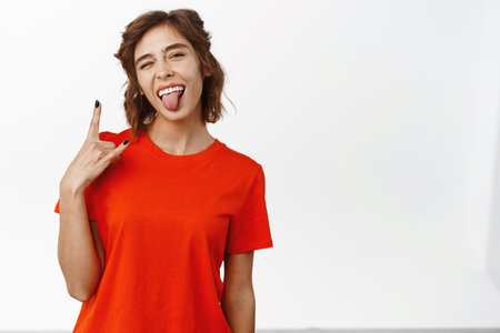 Portrait of enthusiastic young woman having fun, showing rock on, heavy metal sign and tongue, enjoying concert, music festival, standing over white backgroundの写真素材