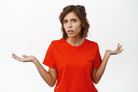 Image of confused brunette girl shrugging and looking clueless, cant understand smth, dont know, standing in red tshirt over white backgroundの写真素材