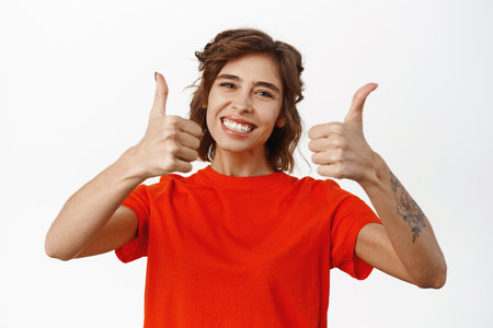 Smiling young woman showing thumbs up, approve or recommend something good, praise nice job, standing over white backgroundの写真素材