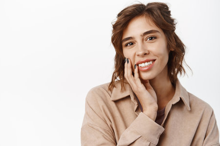 Beauty. Feminine young woman with curly short hairstyle, white smile, natural clear skin without blemishes or acne, touching her face and looking happy, standing over white backgroundの写真素材