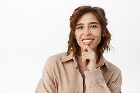 Beauty. Feminine young woman with curly short hairstyle, white smile, natural clear skin without blemishes or acne, touching her face and looking happy, standing over white backgroundの写真素材