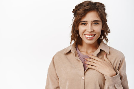 Portrait of smiling girl hold hand on heart and looking pleased, receive praise, talking about personal achievement, standing over white backgroundの写真素材