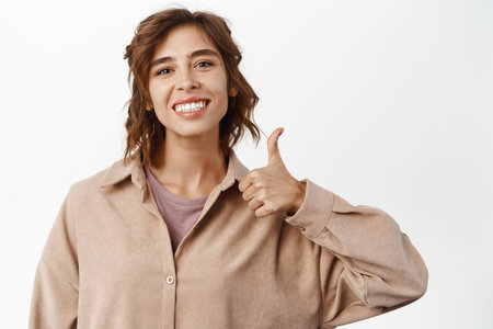 Close up portrait of young woman showing like, thumb up gesture, approve, recommend quality product, standing against white studio backgroundの写真素材