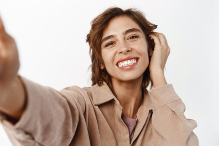Close up portrait of smiling brunette girl taking selfie, touching her face, posing for photo, picture of herself on mobile phone, standing over white backgroundの写真素材