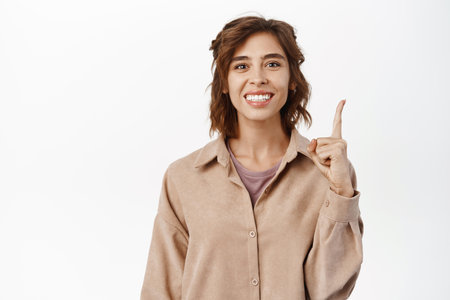 Portrait of smiling brunette girl with curly hairstyle, pointing finger up, showing announcement, promo text on top, demonstrating, standing against white backgroundの写真素材