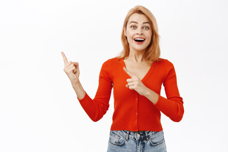 Portrait of smiling redhead woman, looking with excitement and happiness, pointing finger left, showing advertisement or promo logo, standing over white backgroundの写真素材