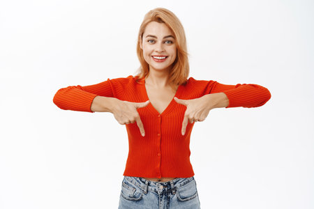 Excited young woman pointing fingers down at logo or banner. Girl showing information below, standing over white backgroundの写真素材