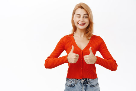 Smiling beautiful woman shows thumbs up in approval. Girl likes and approves smth, stands over white backgroundの写真素材