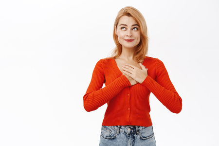 Beautiful smiling redhead woman waiting with excitement for something, holding hand on heart and daydreaming, looking hopeful, standing over white backgroundの写真素材