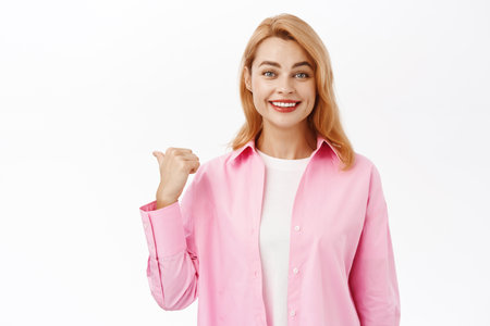 Portrait of redhead girl points finger left, shows advertisement, website logo or product banner, stands in casual clothes over white backgroundの写真素材