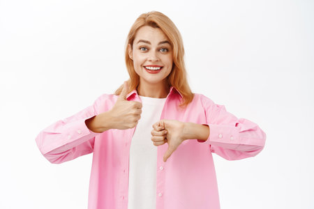 Good or bad. Smiling redhead woman showing thumbs up thumbs down, like or dislike, making decision, standing over white backgroundの写真素材