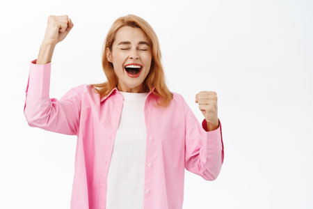 Portrait of excited happy woman, chanting and celebrating, raising fists up and scream of joy, triumphing, standing over white backgroundの写真素材