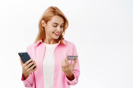 Portrait of smiling woman using credit card and mobile phone app for online shopping, order on website, standing over white backgroundの写真素材
