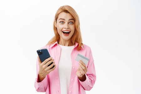 Mobile shopping, online application concept. Smiling young woman holding smartphone and credit card, looking excited, standing over white backgroundの写真素材
