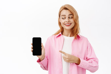 Portrait of smiling young woman showing mobile screen, smartphone app, standing over white studio backgroundの写真素材
