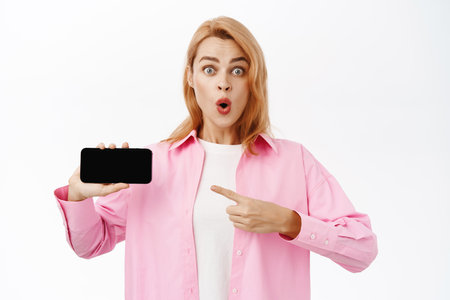 Impressed girl showing mobile phone horizontal screen, looking amazed, standing over white backgroundの写真素材