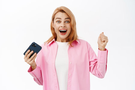 Excited young woman rejoicing, raising hand up and celebrating, holding smartphone, receive good news over mobile phone notification, standing over white backgroundの写真素材