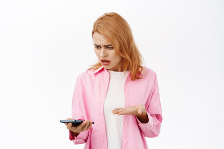 Young woman facing problem on mobile phone, looking frustrated and upset at smartphone, standing over white backgroundの写真素材