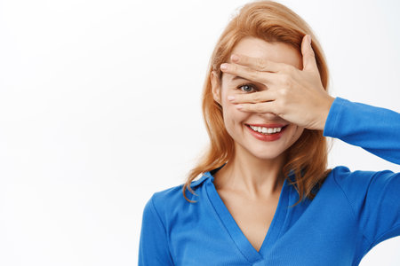 Close up portrait of smiling happy woman covers her eyes, peeks through fingers, intrigued by surprise, stands over white backgroundの写真素材