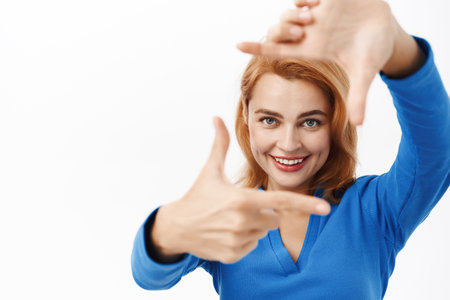 Close up portrait of smiling creative woman, looks through hand frames, imaging, picturing smth, measuring angle of photo, stands over white backgroundの写真素材