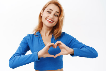 Close up portrait of natural young woman, shows heart sign and looks caring at camera, I love you gesture, stands over white backgroundの写真素材