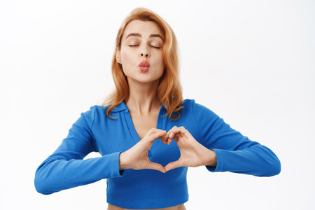 Cute romantic woman shows heart sign, kissing with closed eyes, express her care and affection, stands over white studio backgroundの写真素材