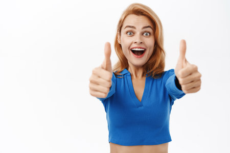 Portrait of happy woman showing thumbs up with amazed face, say yes, approve smth awesome, stands over white backgroundの写真素材