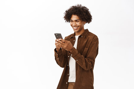 Image of young hispanic man holding mobile phone, using smartphone app for chat, standing over white backgroundの写真素材
