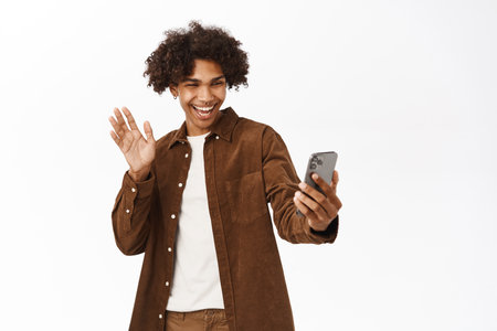 Portrait of handsome boy video chat, talking on mobile phone application, standing over white background, waving hand at smartphoneの写真素材
