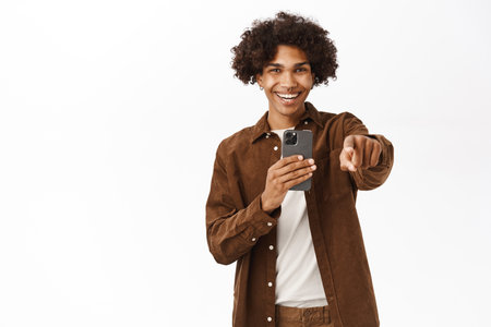 Portrait of gen-z guy pointing at camera, recording video or taking photo on smartphone camera, smiling and laughing, standing over white backgroundの写真素材