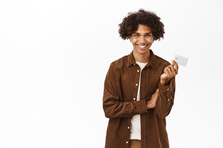 Young hispanic guy holds his credit card, smiles and looks pleased by bank service, stands over white backgroundの写真素材