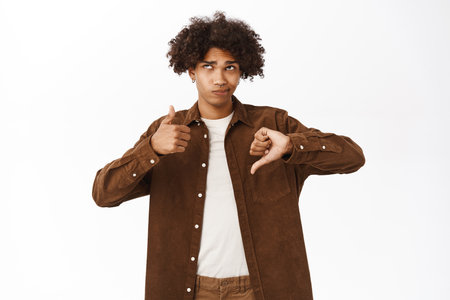Decision, weighing concept. Young man with doubts showing thumbs up and thumbs down, like or dislike, standing over white backgroundの写真素材