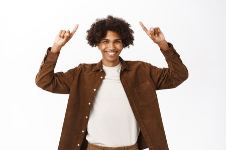 Portrait of smiling hispanic young man showing announcement on top, pointing fingers up and laughing happily, standing over white backgroundの写真素材