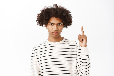 Close up of upset young boy pointing finger up, frowning and looking annoyed, showing smth upsetting, standing over white backgroundの写真素材