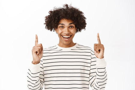 Close up portrait of excited young guy pointing fingers up, showing advertisement and smiling, standing over white backgroundの写真素材